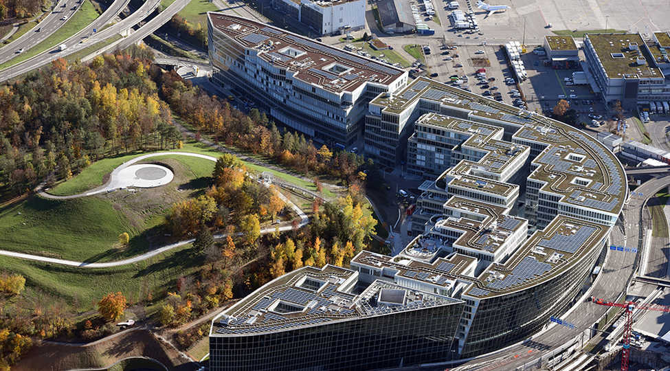 Luftaufnahme von The Circle am Flughafen Zürich. Kreisrunde Gebäudeanordnung, in der Mitte parkähnliche Anlage mit viel Grün.
