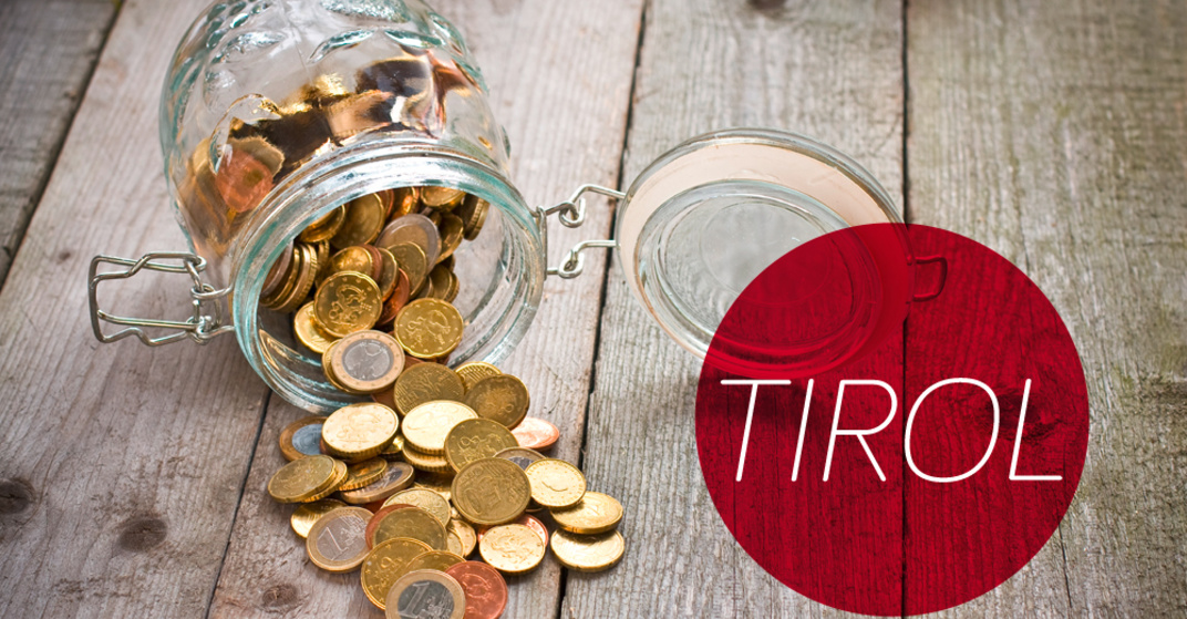 Gekipptes Einmachglas mit verschiedenen Euro- und Centmünzen liegt umgekippt auf einem rustikalen Holzboden, dazu der Text "TIROL" in rotem Kreis.
