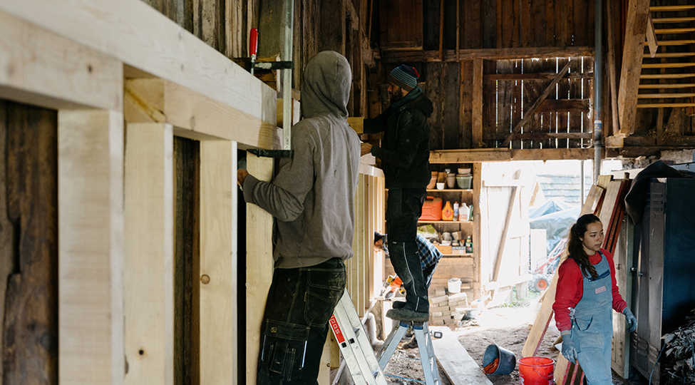 2 Studenten von BASEhabitat stehen auf Leitern und fixieren eine Holzbalkenkonstruktion. Studentin in blauer Latzhose und rotem Pulli geht durch den Raum.