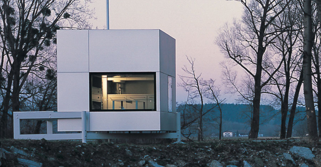 Ein micro-compact Home, entwickelt in Oberösterreich, steht an einem Flussufer im Sonnenuntergang, Blick durchs Fenster ins beleuchtete Innere